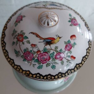 May include: A white porcelain serving dish with a lid featuring a colorful bird and floral design. The lid has a gold knob and a black and gold patterned border. The dish is light blue.