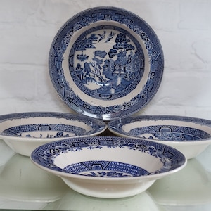 May include: Four white and blue Willow pattern bowls. The bowls feature a blue rim and a detailed blue landscape scene in the centre. The bowls are arranged on a reflective surface against a white brick wall.