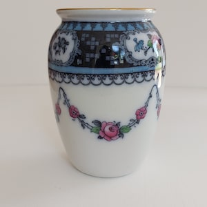 May include: A white ceramic vase with a gold rim and floral design. The vase features a blue and black band at the top with ornate detailing. A garland of pink roses and green leaves decorates the body of the vase.