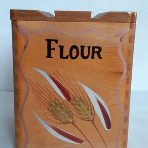 Puede incluir: Contenedor de harina de madera con tapa. La parte frontal del contenedor est&aacute; pintada con la palabra "FLOUR" en negro y espigas de trigo en dorado, rojo y blanco. El contenedor es de color marr&oacute;n claro con una tapa marr&oacute;n m&aacute;s oscura.