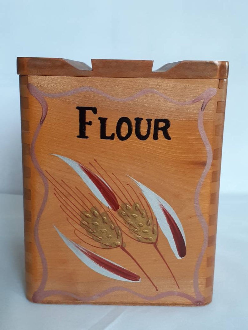 Puede incluir: Contenedor de harina de madera con tapa. La parte frontal del contenedor est&aacute; pintada con la palabra "FLOUR" en negro y espigas de trigo en dorado, rojo y blanco. El contenedor es de color marr&oacute;n claro con una tapa marr&oacute;n m&aacute;s oscura.
