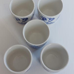 May include: Five white ceramic cups with a blue bird and floral design. The cups are cylindrical with a slightly flared rim. The blue design wraps around the lower portion of each cup. The cups are arranged in a circular pattern.
