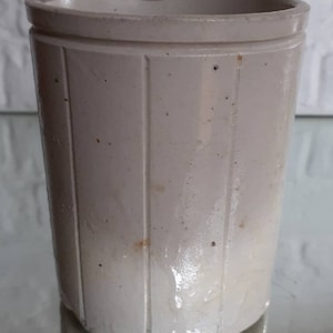 May include: A light beige, cylindrical stoneware jar with vertical grooved detailing. The jar has a slightly flared rim and a smooth, reflective surface. The antique-style container is likely for kitchen or home decor.
