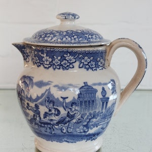 May include: A vintage white and blue ceramic creamer with a lid. The creamer features a detailed blue transferware design of classical figures and architecture. The rim and lid are decorated with a floral pattern. The handle is a light beige color.