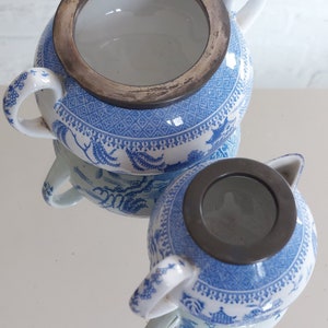 May include: Two blue and white porcelain sugar bowls with silver-colored rims. The bowls have a traditional Asian-inspired design with trees and buildings. The handles are white with blue floral accents. The bowls are reflected on a mirrored surface.