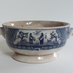 May include: A cream-colored, footed bowl with blue and white Asian-inspired illustrations. The bowl features a detailed landscape scene and decorative handles. The rim and interior have a light brown hue.