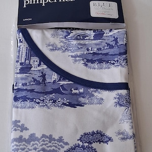 May include: A white apron with a blue Italian landscape design. The apron has a dark blue trim and is packaged in a clear plastic bag. The packaging includes the brand name "Pimpernel" and the words "Blue Italian" and "Spode design".