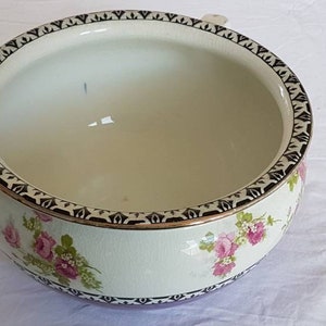 May include: A round, off-white ceramic bowl with a floral design. The bowl features pink roses and green leaves, with a black and gold geometric border. The interior is a light cream color.