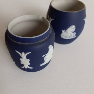 May include: Two small, dark blue ceramic jars with white relief designs. The jars have a rounded shape with a white interior and a matte finish. The relief designs include classical figures and floral motifs.