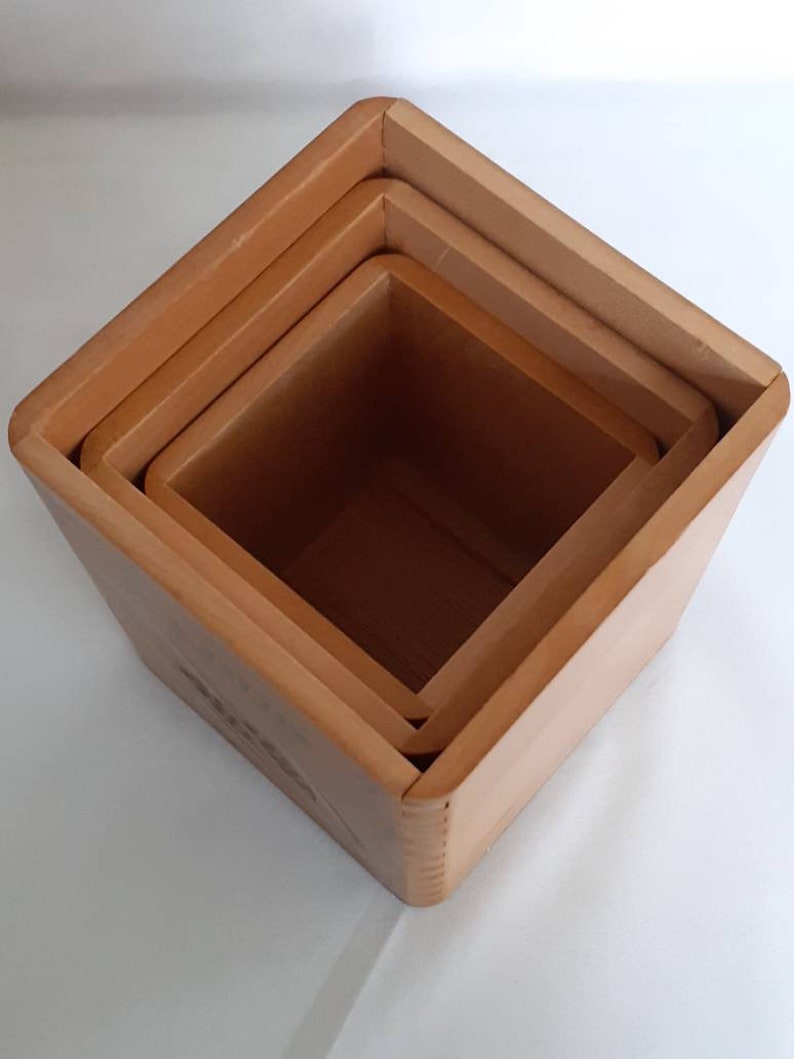 Puede incluir: Juego de cuatro cajas anidadas cuadradas de madera de diferentes tama&ntilde;os. Las cajas son de color marr&oacute;n claro y tienen un dise&ntilde;o sencillo. Las cajas est&aacute;n apiladas unas dentro de otras.