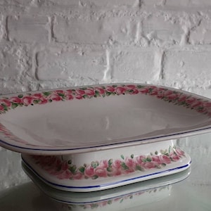 May include: White ceramic serving dish with a raised base, decorated with pink roses and green leaves around the rim and base. A thin blue line borders the rim. The dish is rectangular with rounded corners.