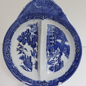 May include: A blue and white divided serving dish with a handle. The dish features a detailed, traditional pattern with scenes of buildings, trees, and floral elements. The rim and handle have a dotted design.