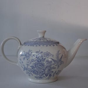 May include: White ceramic teapot with a lid, featuring a blue floral pattern. The teapot has a curved handle and a spout. The design includes flowers and birds. The teapot is a classic design.