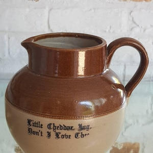 May include: A brown and tan ceramic jug with a handle and spout. The jug has a rounded body and a darker brown top. The text "Little Cheddar Jug, Don't I Love Thee" is printed on the jug.