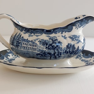 Könnte beinhalten: Cremefarbene Sauciere und passende Untertasse mit blauem Transferdruck. Das Muster zeigt eine Landschaft mit einem Herrenhaus und Bäumen. Die Sauciere hat einen gebogenen Griff und einen gewellten Rand.