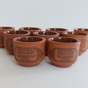 May include: A set of ten terracotta egg cups. Each cup has a brown interior and a label with the text "THE ORIGINAL EGG CUP" and "HENRY WATSON POTTERY ENGLAND Founded 1800". The cups are arranged in a group.