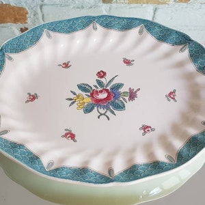 May include: Oval serving platter with a scalloped edge and a floral design. The platter has a light pink center with a central bouquet of red, yellow, and blue flowers. The border is teal with a scale pattern.
