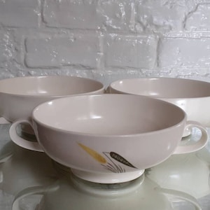 Puede incluir: Tres cuencos de sopa de cerámica color crema con asas. Un cuenco presenta un diseño decorativo con detalles amarillos y negros. Los cuencos están sobre una superficie reflectante, con un fondo de ladrillos blancos.