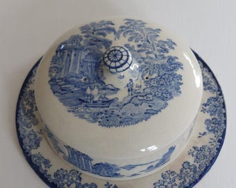 May include: A blue and white ceramic butter dish with a lid featuring a scenic landscape design. The dish sits on a matching plate with a floral border. The lid has a small, decorative knob.