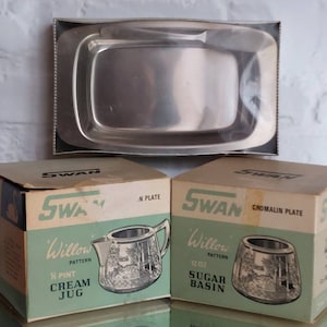 Puede incluir: Una mantequera de acero inoxidable con un cuchillo y dos cajas de vajilla Swan Willow Pattern. Una caja está etiquetada como "½ Pint Cream Jug" y la otra como "12 oz Sugar Basin". Las cajas tienen un esquema de color verde y beige.
