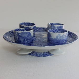 May include: A blue and white ceramic serving tray with four matching cups. The tray has a raised pedestal base and a detailed landscape pattern. The cups and tray feature a classic blue and white color scheme, perfect for serving tea or small treats.