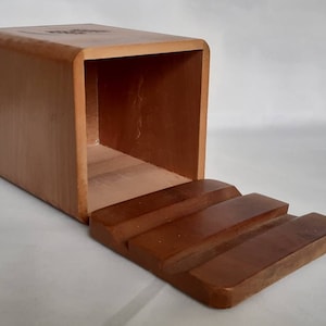 Puede incluir: Una caja rectangular de madera con una parte superior curvada y una tapa con bisagras escalonada. La caja es de color marr&oacute;n claro y la tapa es de un tono marr&oacute;n m&aacute;s oscuro. La caja parece estar vac&iacute;a.