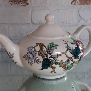 May include: Cream-colored ceramic teapot with a lid and handle. The teapot is decorated with a floral and botanical design featuring blue, green, and red flowers and leaves, and brown branches. The teapot is sitting on a reflective surface.