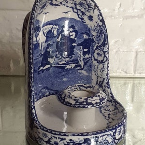 May include: Blue and white ceramic wall-mounted candle holder. The candle holder features a detailed pastoral scene and floral patterns. The design includes a small candle cup and a curved back, with a reflective surface below.