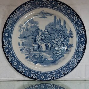 May include: A blue and white decorative plate with a detailed landscape scene. The plate features a central image of figures in a garden setting, surrounded by a patterned border. The plate is likely ceramic and has a vintage aesthetic.