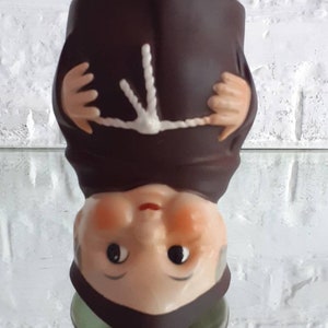 May include: A ceramic figurine of a monk, upside down. The figure is dressed in a brown robe with a white rope belt and a dark brown hood. The face is light-skinned with black eyes, a red mouth, and rosy cheeks.