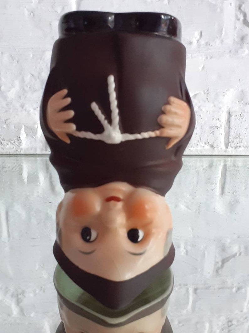 May include: A ceramic figurine of a monk, upside down. The figure is dressed in a brown robe with a white rope belt and a dark brown hood. The face is light-skinned with black eyes, a red mouth, and rosy cheeks.