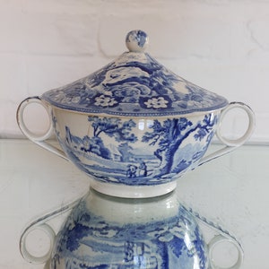 May include: A blue and white ceramic soup tureen with a lid. The tureen features a landscape design with trees, buildings, and figures. It has two handles and a small knob on the lid. The tureen is reflected on a glass surface.