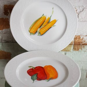 May include: Two white ceramic plates with a textured border. One plate features a vibrant illustration of three ears of corn. The other plate displays a red and orange bell pepper illustration. A decorative kitchenware set.