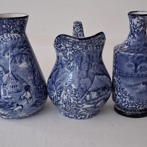 May include: Three blue and white ceramic pieces with a pastoral scene design. The set includes a vase, a small pitcher, and a bottle. The pieces have a detailed, vintage aesthetic, with a dark blue and white color scheme.