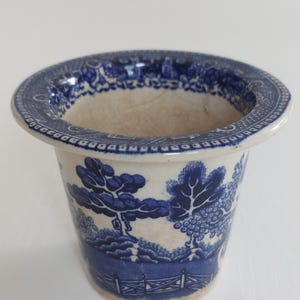May include: A blue and white ceramic planter with a classic Willow pattern design. The planter features a detailed landscape scene with trees, a fence, and a bridge. The rim has a decorative border.