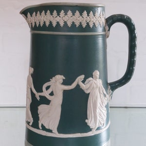 May include: A teal-colored ceramic pitcher with a white relief design of dancing figures. The pitcher has a handle and a decorative rim with a white leaf pattern. The base and rim are accented with a light gray trim.