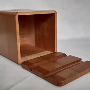 Puede incluir: Caja de madera rectangular con tapa con bisagras. La caja es de color marr&oacute;n claro con esquinas redondeadas. La tapa, de un tono m&aacute;s oscuro, presenta un dise&ntilde;o escalonado. El interior de la caja es visible.