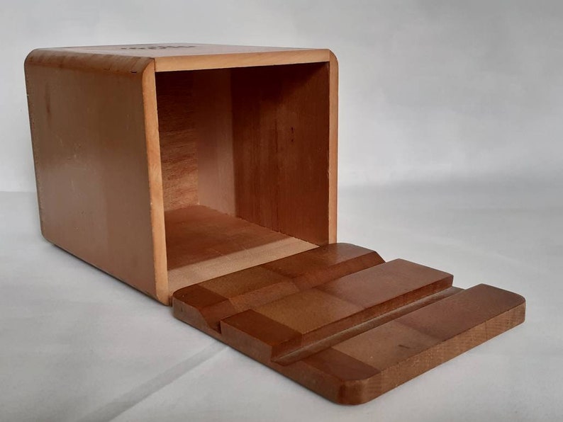 Puede incluir: Caja de madera rectangular con tapa con bisagras. La caja es de color marr&oacute;n claro con esquinas redondeadas. La tapa, de un tono m&aacute;s oscuro, presenta un dise&ntilde;o escalonado. El interior de la caja es visible.