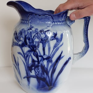 May include: A white and blue ceramic pitcher with a floral iris design. The pitcher has a curved spout, a decorative handle, and a blue rim. The iris design is in shades of blue.