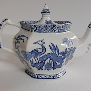 May include: White ceramic teapot with blue floral and bird design. The teapot features a hexagonal body, a curved handle, and a spout. The lid is topped with a finial. The blue design includes a bird, flowers, and geometric patterns.