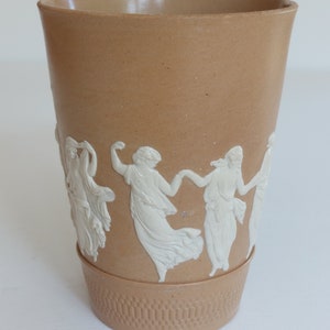 May include: Tan ceramic vase with a textured base and a band of white relief figures dancing around the middle. The vase has a slightly flared top and a smooth, unadorned interior.