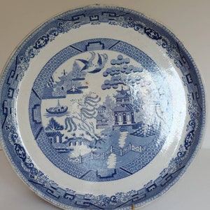 May include: A large, round, blue and white ceramic plate with a Willow pattern design. The plate features a central scene of a bridge, buildings, and birds, surrounded by a decorative border. The plate is approximately 30 cm in diameter.