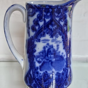 May include: A blue and white ceramic pitcher with a handle and spout. The pitcher features a detailed landscape scene with a domed building, trees, and floral patterns. The design is in a classic blue and white style, with a glossy finish.