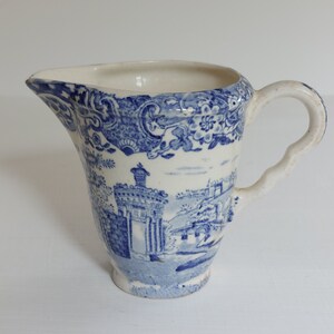 May include: Antique blue and white ceramic pitcher with a landscape design. The pitcher features a spout, handle, and a detailed pattern of buildings and trees. The pitcher is approximately 6 inches tall.