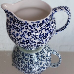 May include: A small, round creamer with a handle and spout. The creamer is white on the inside and covered in a blue floral pattern on the outside. The creamer is reflected in the surface below.
