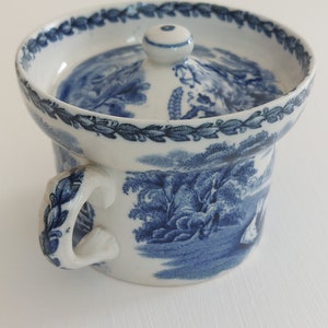 May include: A white and blue ceramic chamber pot with a lid. The pot features a handle and a detailed landscape design in blue. The rim and handle are decorated with blue floral patterns. The lid has a small knob.