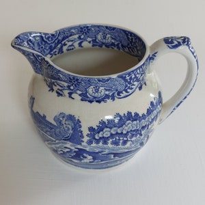 May include: A white ceramic pitcher with a blue floral and landscape design. The pitcher has a spout and a handle. The rim and handle are decorated with blue floral patterns. The body of the pitcher features a blue landscape scene.