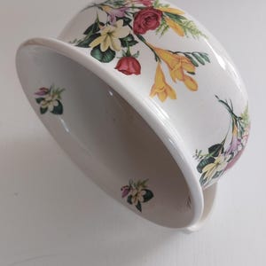 May include: White ceramic bowl with a floral design featuring red roses, yellow lilies, and other colorful flowers. The bowl has a wide rim and a curved shape, suitable for serving or display.