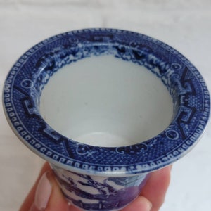 May include: A small, white ceramic cup with a blue Willow pattern design. The cup has a wide, flared rim with a detailed border. The blue and white design features a landscape scene. The cup is held by a person.