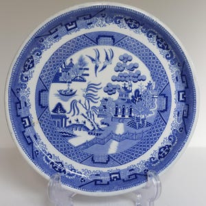 May include: A round, blue and white Willow pattern plate. The plate features a detailed landscape scene with a bridge, buildings, trees, and birds. The border has a geometric design. The plate is displayed on a clear plastic stand.